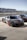 A sleek sports car with a black carbon fiber finish and red accents is parked on a concrete surface next to other similar vehicles. The setting appears to be a race track pit stop with barriers and spectator stands visible in the background under a clear blue sky.