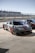 A sleek sports car with a black carbon fiber finish and red accents is parked on a concrete surface next to other similar vehicles. The setting appears to be a race track pit stop with barriers and spectator stands visible in the background under a clear blue sky.