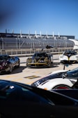 A group of high-end sports cars parked at a racetrack, with one black car prominently in front with its doors open upwards. The background features a racing facility, complete with grandstands and a metal fence. The sky is clear and blue, contributing to the overall sleek and futuristic ambiance.