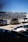 A group of high-end sports cars parked at a racetrack, with one black car prominently in front with its doors open upwards. The background features a racing facility, complete with grandstands and a metal fence. The sky is clear and blue, contributing to the overall sleek and futuristic ambiance.
