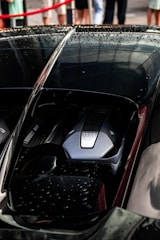 A close-up of a sleek car engine with the number 1500 prominently displayed. The car's exterior is black, shiny, and wet from rain, with droplets visible on its surface. In the background, there are blurred figures of people and a red barricade rope.