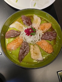 A round plate with an arrangement of mixed sashimi pieces, including salmon, tuna, and other fish, beautifully presented in a circular pattern. At the center, there is a garnish of shredded daikon and a pink flower with green leaves.