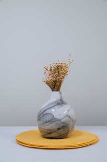A marble-patterned vase filled with dry ornamental flowers sits on a round yellow placemat against a plain light gray background.