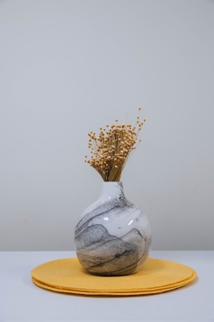 A marble-patterned vase filled with dry ornamental flowers sits on a round yellow placemat against a plain light gray background.