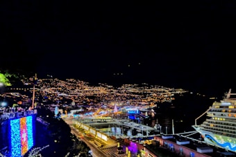 A vibrant nighttime cityscape with a large number of twinkling lights scattered across a hillside, a cruise ship docked at the shoreline, and colorful illuminations adorning buildings and streets near a harbor.