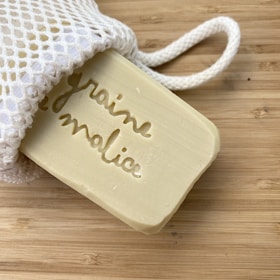 A beige bar of soap with an engraving partially visible emerges from a white mesh bag, placed on a light wooden surface.