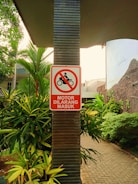 A sign with a red border and a symbol of a motorcycle inside a red circle with a diagonal line through it is mounted on a vertical pillar. Below the symbol, the text 'MOTOR DILARANG MASUK' is written, indicating motorcycles are not allowed to enter. The surrounding area includes lush green foliage and a paved pathway.