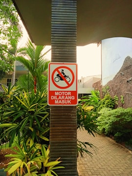 A sign with a red border and a symbol of a motorcycle inside a red circle with a diagonal line through it is mounted on a vertical pillar. Below the symbol, the text 'MOTOR DILARANG MASUK' is written, indicating motorcycles are not allowed to enter. The surrounding area includes lush green foliage and a paved pathway.