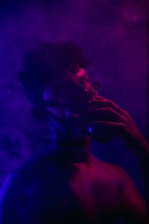 A person wearing a gas mask is immersed in a dark, atmospheric setting filled with purple and blue smoke. The lighting casts deep shadows and highlights the contours of the figure's face and body, creating a mysterious and enigmatic ambiance.