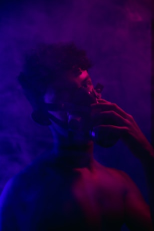 A person wearing a gas mask is immersed in a dark, atmospheric setting filled with purple and blue smoke. The lighting casts deep shadows and highlights the contours of the figure's face and body, creating a mysterious and enigmatic ambiance.