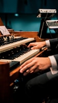 A person is playing an organ keyboard with both hands, surrounded by additional music equipment. The setting appears to be a dimly lit venue or studio.