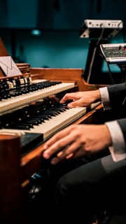 A person is playing an organ keyboard with both hands, surrounded by additional music equipment. The setting appears to be a dimly lit venue or studio.
