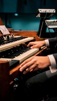 A person is playing an organ keyboard with both hands, surrounded by additional music equipment. The setting appears to be a dimly lit venue or studio.