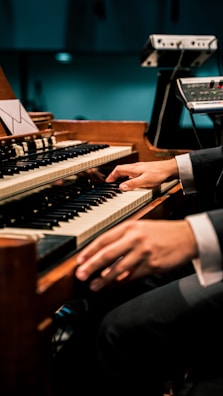 A person is playing an organ keyboard with both hands, surrounded by additional music equipment. The setting appears to be a dimly lit venue or studio.