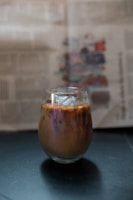 A glass of iced coffee sits on a dark surface with visible ice cubes and a blend of brown and creamy swirls. In the background, there is a blurred newspaper with text and colorful images.