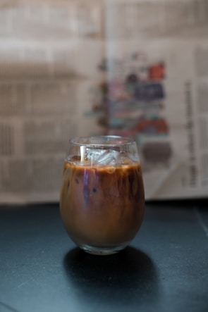 A glass of iced coffee sits on a dark surface with visible ice cubes and a blend of brown and creamy swirls. In the background, there is a blurred newspaper with text and colorful images.