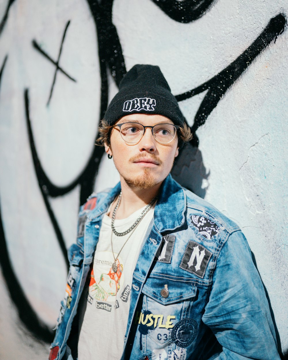 a man wearing glasses and a denim jacket standing in front of a wall with graffiti