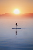 A person paddleboarding on calm water during a serene sunrise, with mountains in the distance and a bright sun low in the sky, creating a peaceful and tranquil scene.