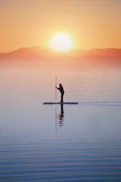 A peaceful paddle boarder gliding on the calm blue lake during sunrise.
