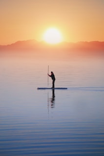 A person paddleboarding on calm water during a serene sunrise, with mountains in the distance and a bright sun low in the sky, creating a peaceful and tranquil scene.