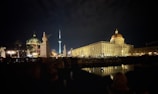 A nighttime cityscape featuring illuminated historic buildings and monuments. A domed structure and a tower are prominent, with reflections visible in the water. Statues and architectural details add to the grandeur of the scene.