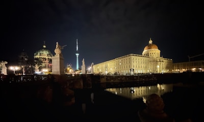 A nighttime cityscape featuring illuminated historic buildings and monuments. A domed structure and a tower are prominent, with reflections visible in the water. Statues and architectural details add to the grandeur of the scene.