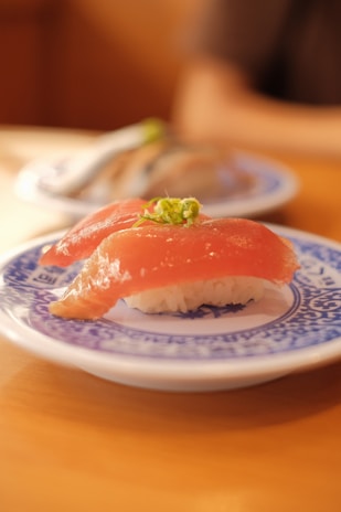 A close-up view of a sushi piece laid on a decorative blue and white ceramic plate. The sushi features a slice of raw fish topped with finely chopped green garnish resting on a bed of white rice. The plate sits on a wooden surface, with another blurred plate of sushi visible in the background, giving a cozy dining ambiance.