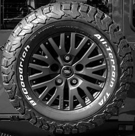 A close-up of a rugged, all-terrain tire mounted on the back of a vehicle featuring bold, deep treads and a multi-spoke wheel design. The brand logo and tire specifications are prominently displayed.