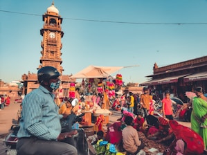 A bustling marketplace with numerous people engaged in various activities. A prominent clock tower stands tall in the background, surrounded by vibrant market stalls selling colorful items. People are seen walking and interacting, with a man wearing a helmet and a face mask on a scooter in the foreground.