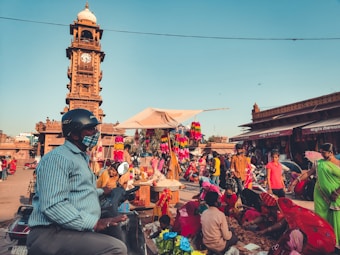 A bustling marketplace with numerous people engaged in various activities. A prominent clock tower stands tall in the background, surrounded by vibrant market stalls selling colorful items. People are seen walking and interacting, with a man wearing a helmet and a face mask on a scooter in the foreground.