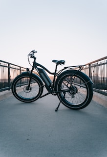 A bicycle is placed on a pathway with railings on both sides. The background shows a clear sky transitioning from blue to a soft orange near the horizon, indicating sunset or sunrise. The bicycle has thick tires and a frame suitable for electric or mountain biking. The setting gives a sense of tranquility and open space.