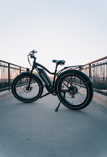 A bicycle is placed on a pathway with railings on both sides. The background shows a clear sky transitioning from blue to a soft orange near the horizon, indicating sunset or sunrise. The bicycle has thick tires and a frame suitable for electric or mountain biking. The setting gives a sense of tranquility and open space.