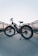 A bicycle is placed on a pathway with railings on both sides. The background shows a clear sky transitioning from blue to a soft orange near the horizon, indicating sunset or sunrise. The bicycle has thick tires and a frame suitable for electric or mountain biking. The setting gives a sense of tranquility and open space.