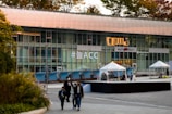 A modern building features a large glass facade displaying the hashtag '#늘ACC' in bold lettering. The foreground includes several people walking, and there are tents set up on a tiled plaza. Trees with autumn-colored leaves are visible on the building's rooftop.