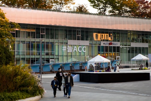 A modern building features a large glass facade displaying the hashtag '#늘ACC' in bold lettering. The foreground includes several people walking, and there are tents set up on a tiled plaza. Trees with autumn-colored leaves are visible on the building's rooftop.