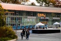 A modern building features a large glass facade displaying the hashtag '#늘ACC' in bold lettering. The foreground includes several people walking, and there are tents set up on a tiled plaza. Trees with autumn-colored leaves are visible on the building's rooftop.