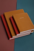Four leather-bound notebooks are neatly stacked on a surface with a geometric color pattern. The top notebook is mustard-colored and labeled 'NOTES' in capital letters, followed by another mustard-colored one, then a red, and finally another red underneath. The background consists of a teal and brown split, adding a stylish and modern feel to the composition.