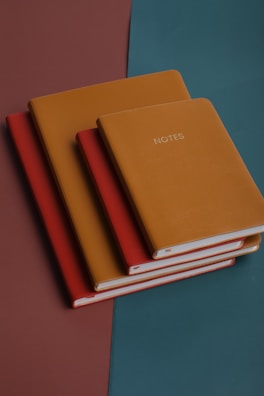 Four leather-bound notebooks are neatly stacked on a surface with a geometric color pattern. The top notebook is mustard-colored and labeled 'NOTES' in capital letters, followed by another mustard-colored one, then a red, and finally another red underneath. The background consists of a teal and brown split, adding a stylish and modern feel to the composition.