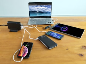 A wooden table holds multiple electronic devices being charged, including a laptop displaying a mountain and ocean scene, a tablet, two smartphones, and a power bank. A charging station is used to connect all devices, with visible charging cables.