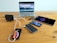 A wooden table holds multiple electronic devices being charged, including a laptop displaying a mountain and ocean scene, a tablet, two smartphones, and a power bank. A charging station is used to connect all devices, with visible charging cables.