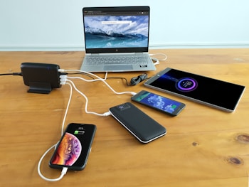 A wooden table holds multiple electronic devices being charged, including a laptop displaying a mountain and ocean scene, a tablet, two smartphones, and a power bank. A charging station is used to connect all devices, with visible charging cables.