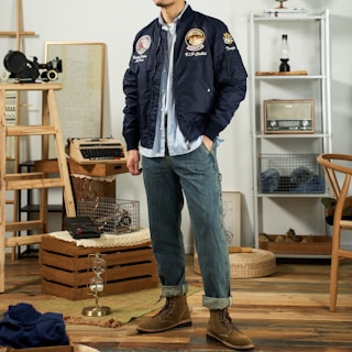 A person is standing in a stylish room, wearing a navy blue bomber jacket with patches, a light blue button-up shirt, and rolled-up denim jeans with brown boots. The background includes various vintage items such as a typewriter, camera, radio, and an hourglass, creating a retro atmosphere.