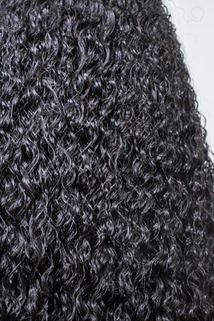 A close-up view of dark, tightly coiled curly hair with a glossy texture. The curls are prominent, well-defined, and densely packed, creating a detailed and uniform pattern.