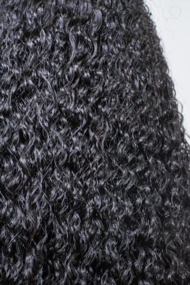 A close-up view of dark, tightly coiled curly hair with a glossy texture. The curls are prominent, well-defined, and densely packed, creating a detailed and uniform pattern.