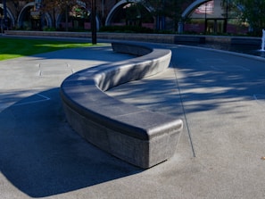 A curved, modern bench made of smooth, dark stone is set in an open plaza area. The surrounding surfaces are paved with gray tiles, and patches of green grass are visible in some areas. In the background, arched windows and buildings with brick and stone facades add to the architectural appeal. The image is taken in daylight with a water fountain visible on the right side.