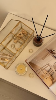 A tabletop features decorative items including a gold-rimmed glass box containing jewelry and trinkets like rings, a crystal, and a pearl necklace. Beside it stands a reed diffuser in a glass container and a lit candle in a glass holder. An open book with interior photography lies nearby.