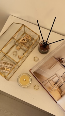 A tabletop features decorative items including a gold-rimmed glass box containing jewelry and trinkets like rings, a crystal, and a pearl necklace. Beside it stands a reed diffuser in a glass container and a lit candle in a glass holder. An open book with interior photography lies nearby.