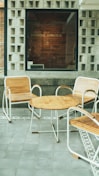 A modern outdoor seating area features three wooden chairs with metal frames together with a round wooden table. The floor is tiled with gray slabs, bordered by a decorative pattern. The background consists of a window with a brick wall inside, where the words 'Byhand Limited' are visible.