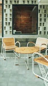 A modern outdoor seating area features three wooden chairs with metal frames together with a round wooden table. The floor is tiled with gray slabs, bordered by a decorative pattern. The background consists of a window with a brick wall inside, where the words 'Byhand Limited' are visible.