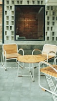 A modern outdoor seating area features three wooden chairs with metal frames together with a round wooden table. The floor is tiled with gray slabs, bordered by a decorative pattern. The background consists of a window with a brick wall inside, where the words 'Byhand Limited' are visible.